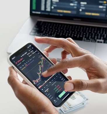 Hands using smartphone trading app with charts, dollar bills in background.